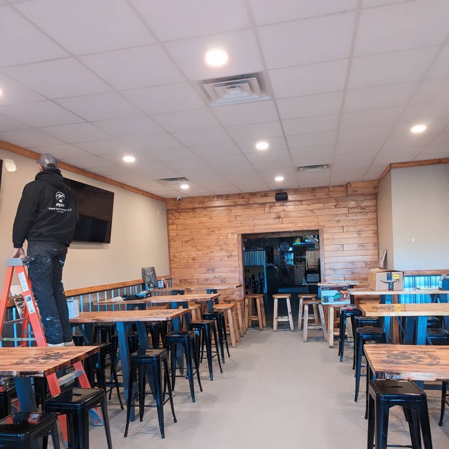 Worker on a ladder installing something in a mostly empty restaurant dining room with wooden tables and stools