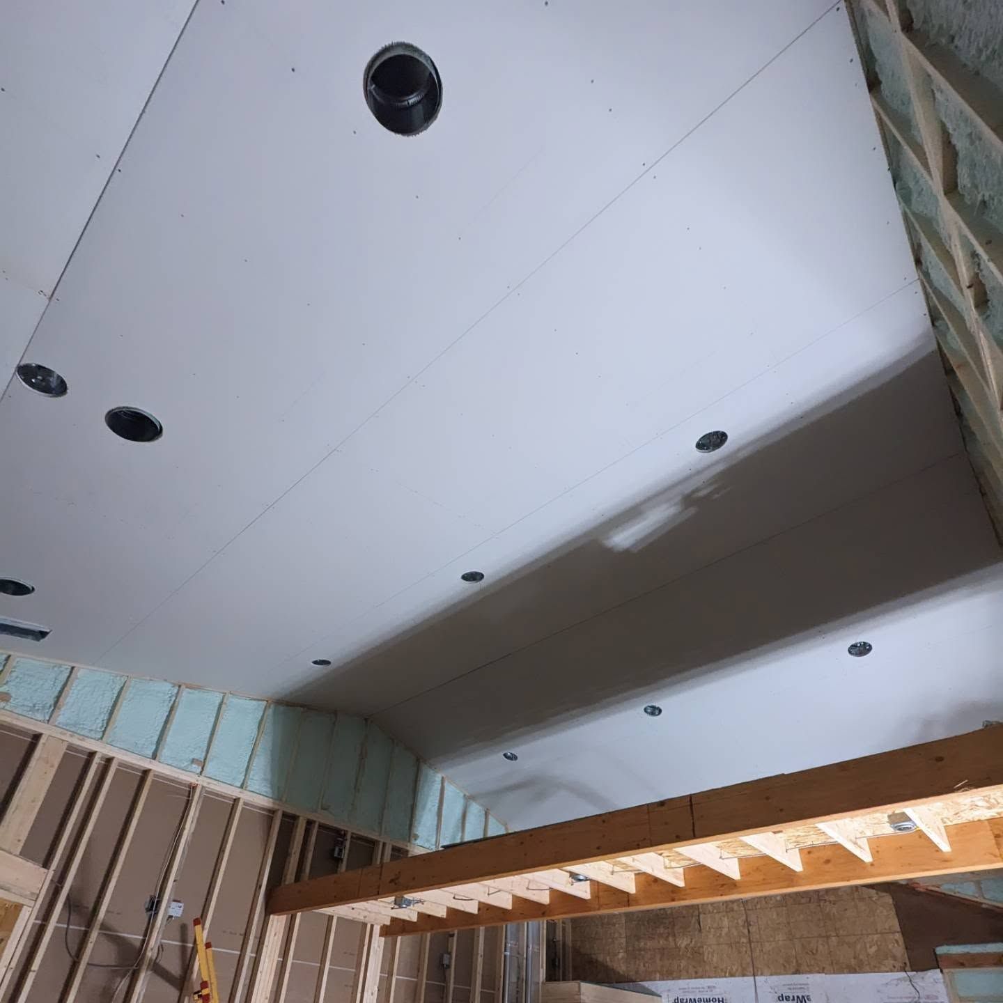 Construction site ceiling with drywall panels, recessed light openings, and exposed framing below.