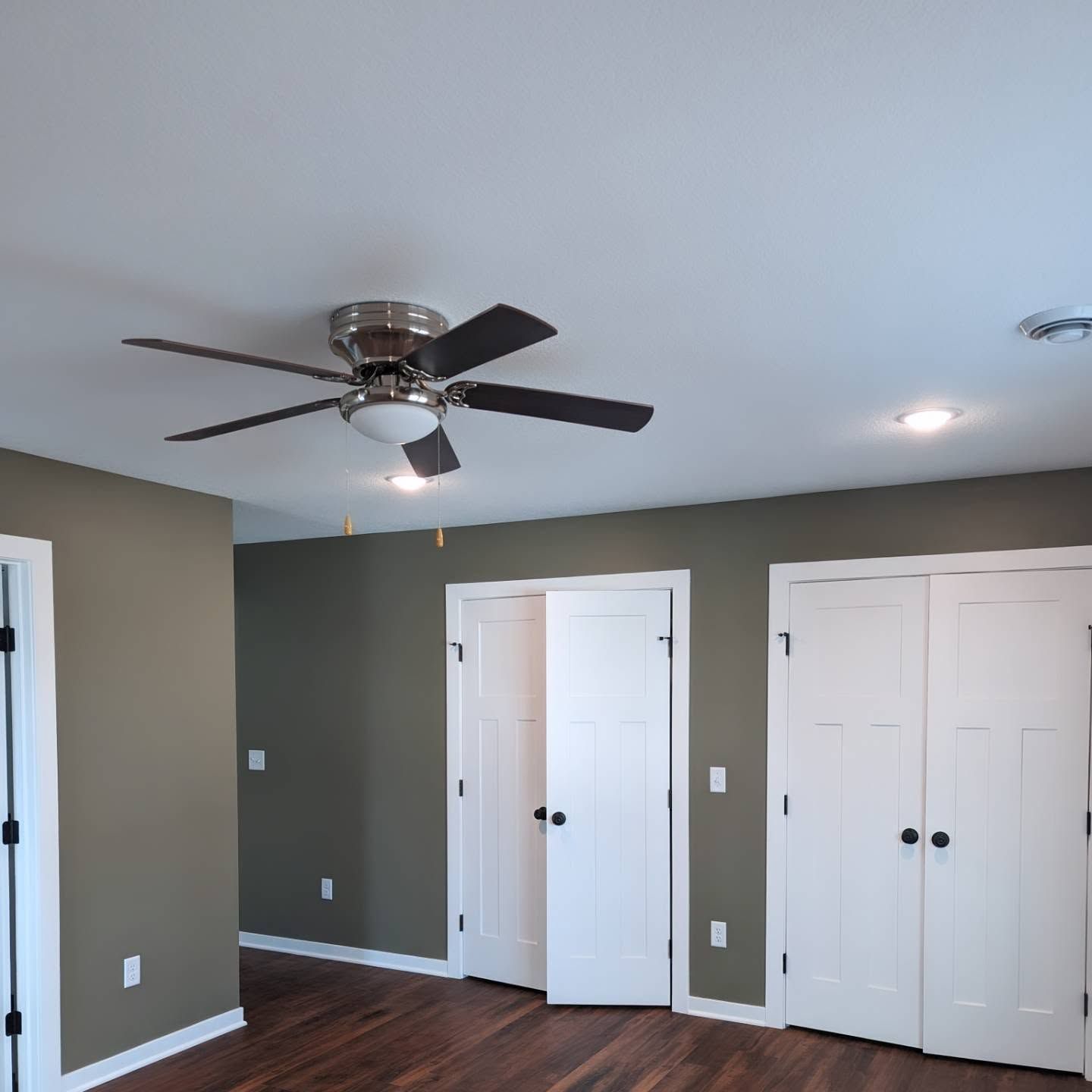 Empty bedroom with gray walls, white closet doors, wood floor, and ceiling fan with light.