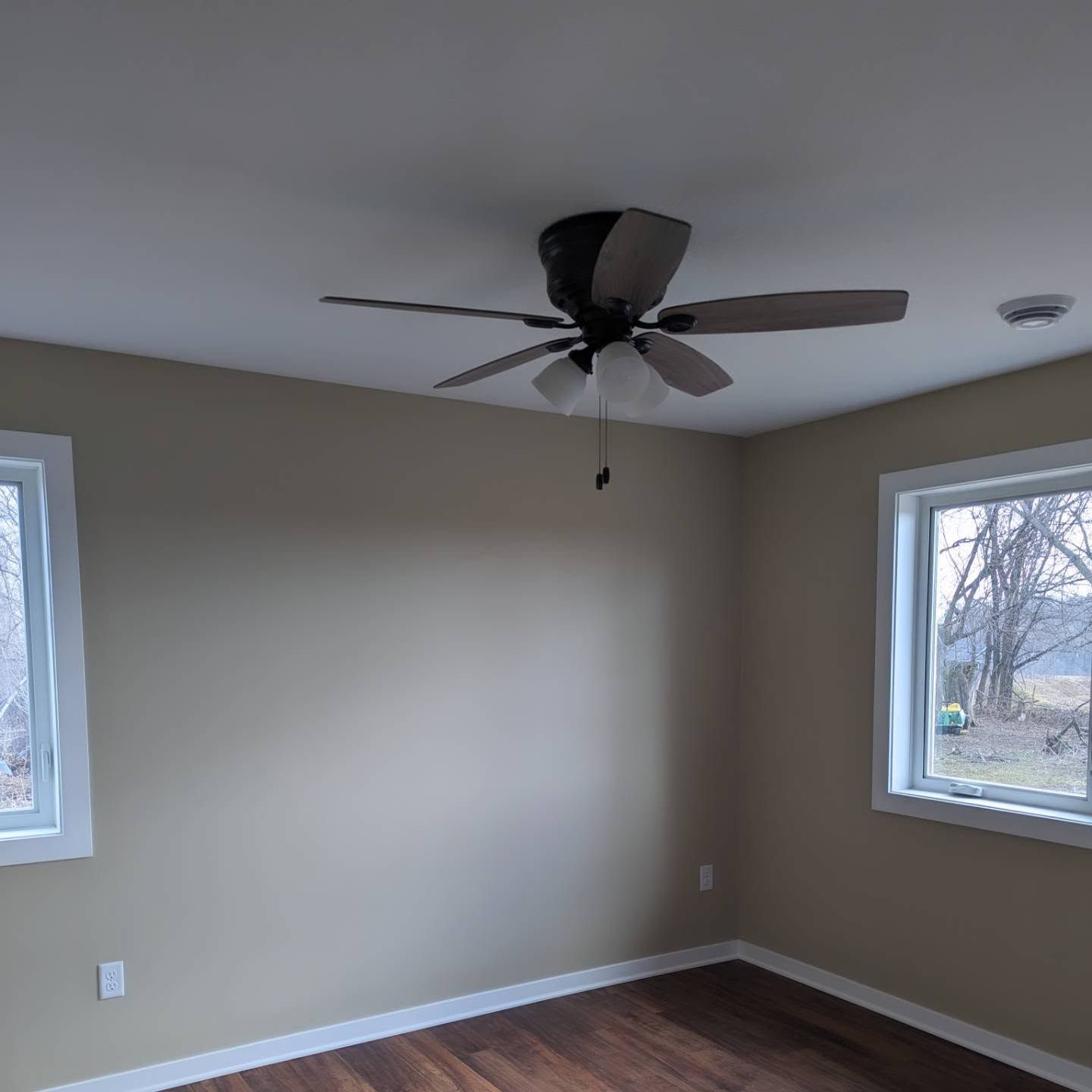 Empty beige room with dark wood floor, two windows, and a ceiling fan.
