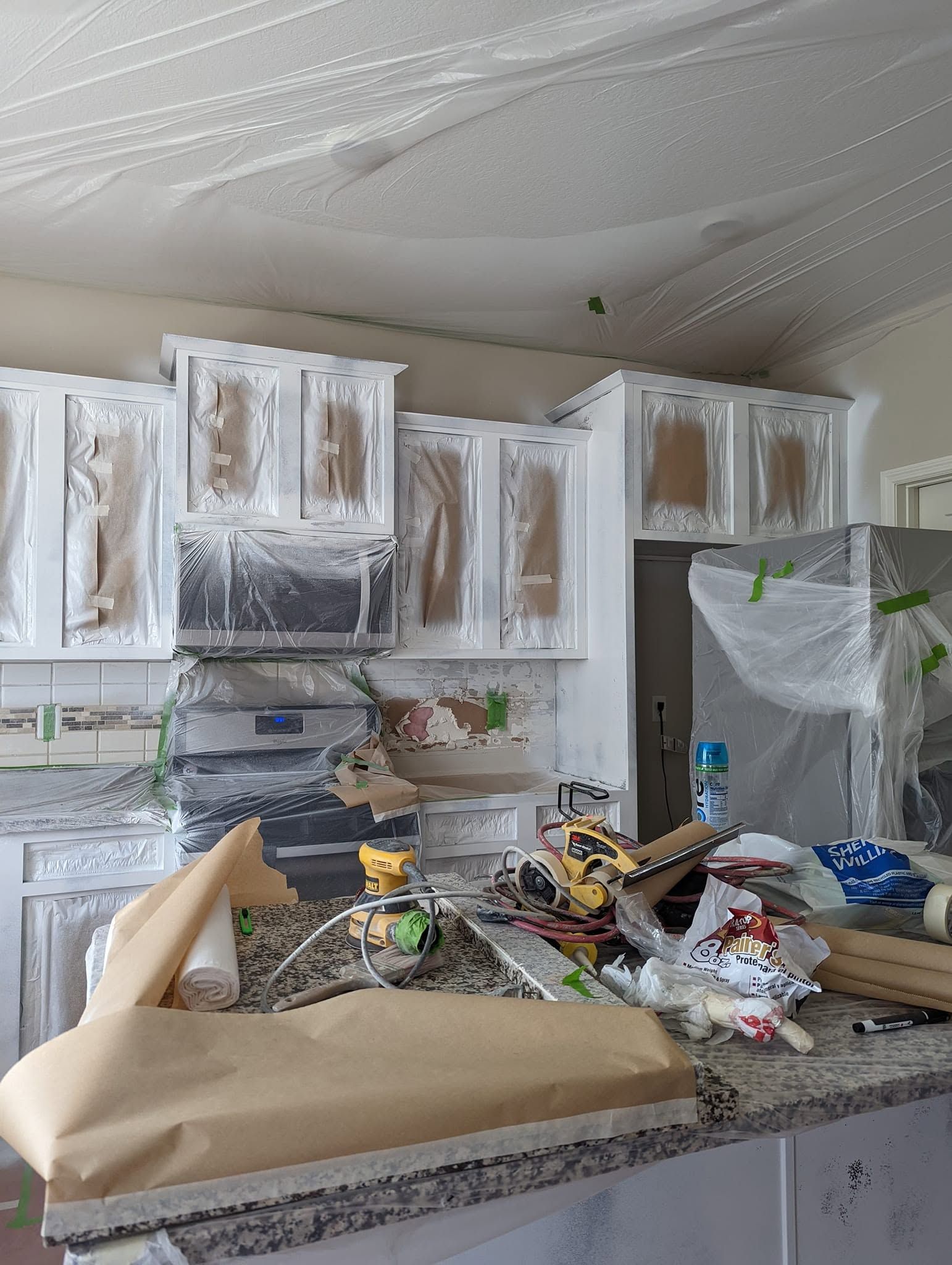 Messy kitchen with white cabinets, plastic-covered appliances, and debris scattered on the countertop.