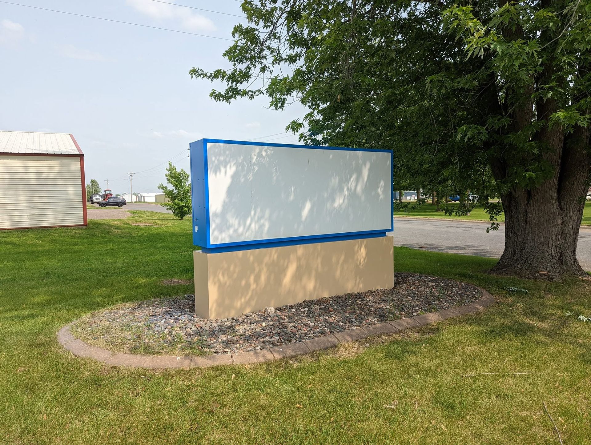 Blank roadside sign with blue border on a grassy verge beside a tree and road