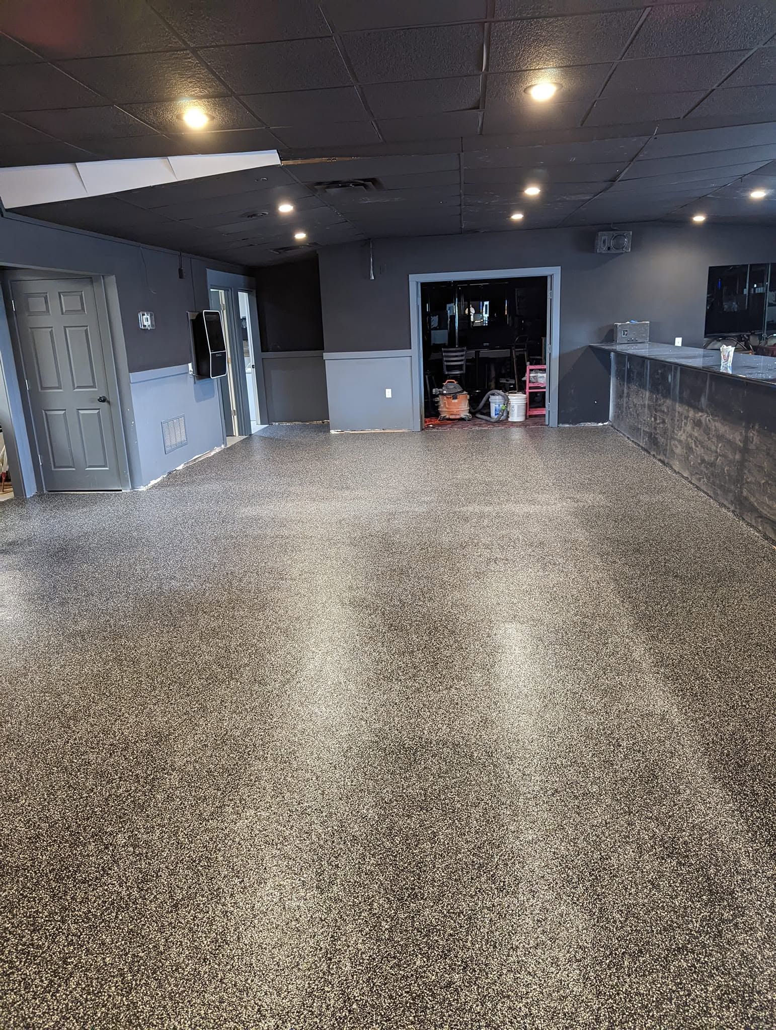Empty indoor room with speckled floor, dark ceiling lights, and an open doorway at the back