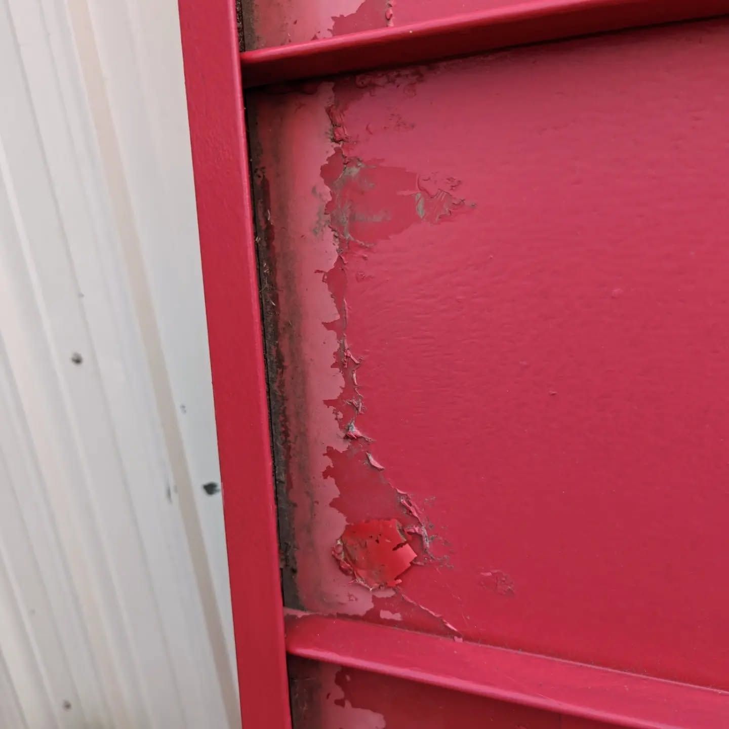 Close-up of red painted metal with peeling paint and rust beside a white corrugated panel