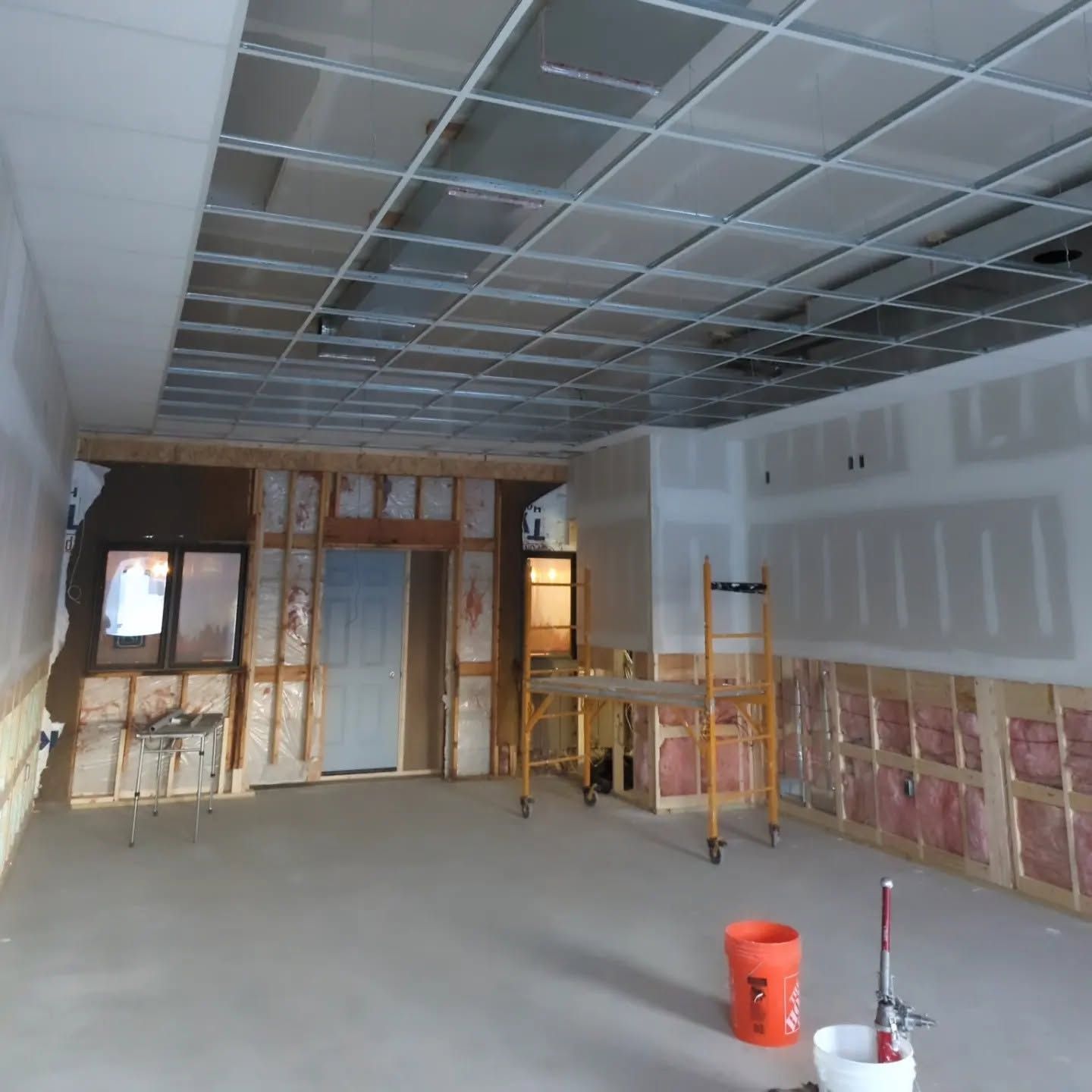 Unfinished room with drywall, exposed ceiling grid, windows, scaffolding, and construction tools on the floor.