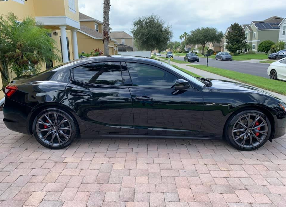 Black Maserati parked on a brick driveway in a suburban neighborhood. Red brake calipers, tinted windows.