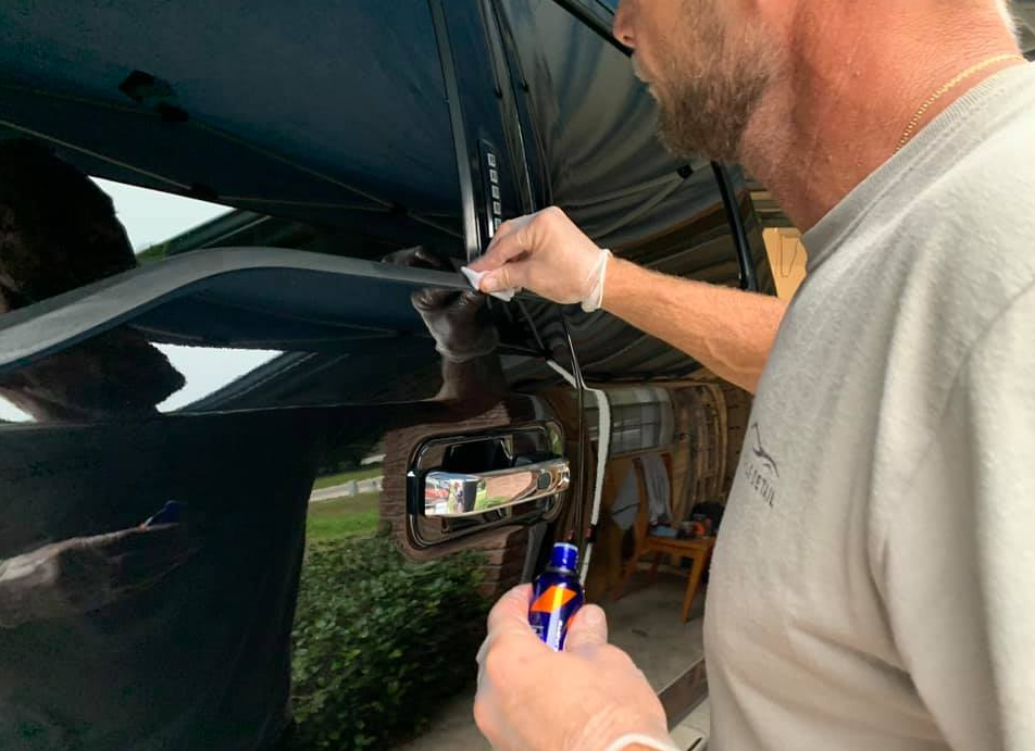 Man in gloves cleaning black car door with a cloth; holding a blue bottle.