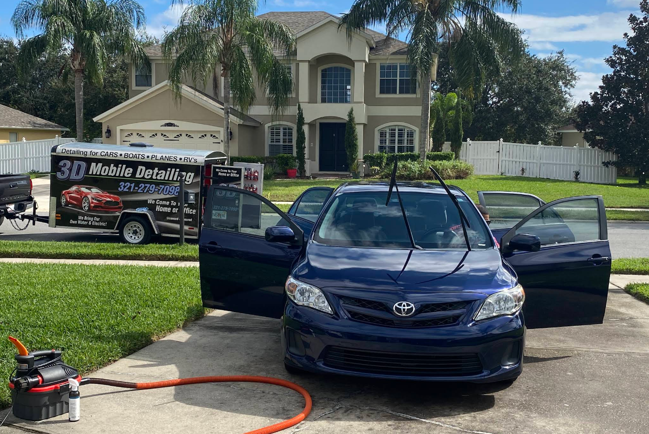 A blue car has open doors as a mobile detailing service cleans it on a driveway in front of a house.