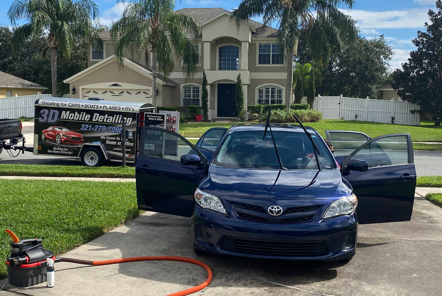 A blue car parked in a driveway being detailed; a mobile detailing trailer and house are in the background.