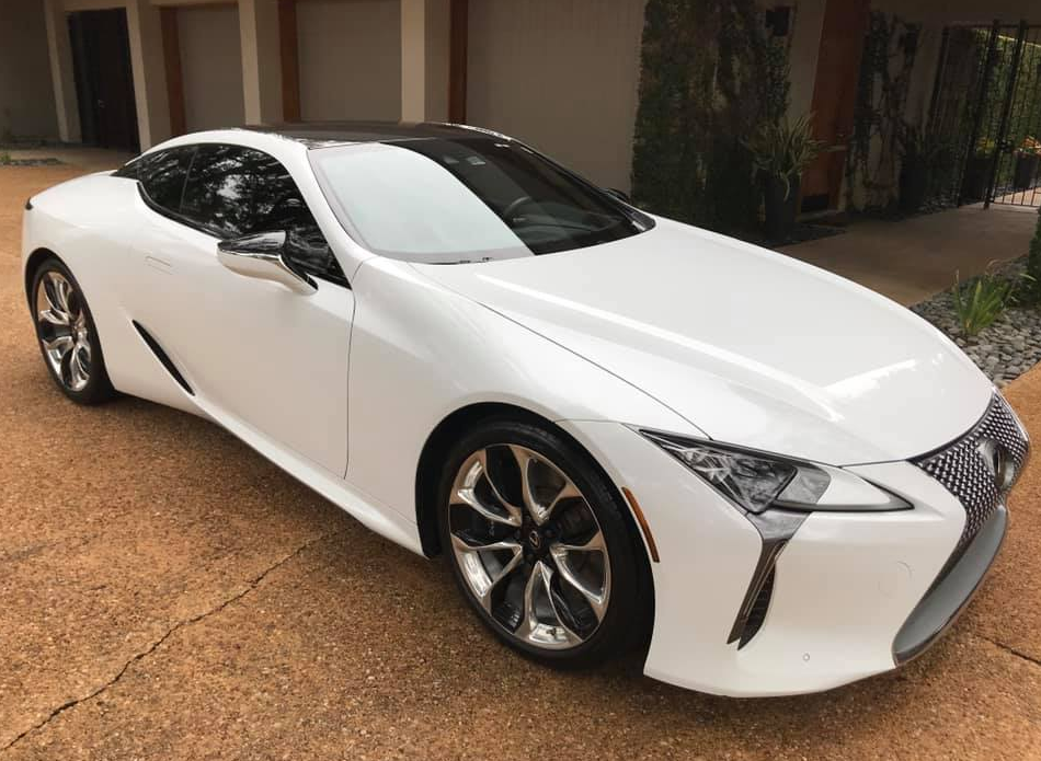 White Lexus LC coupe on a brown brick driveway with black roof, chrome wheels, and chrome side mirrors.