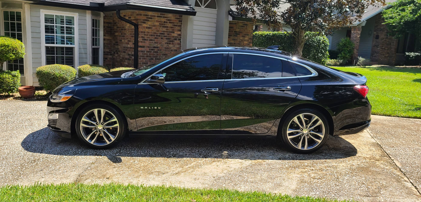 Black car parked in front of a house with a paved driveway and green lawn.