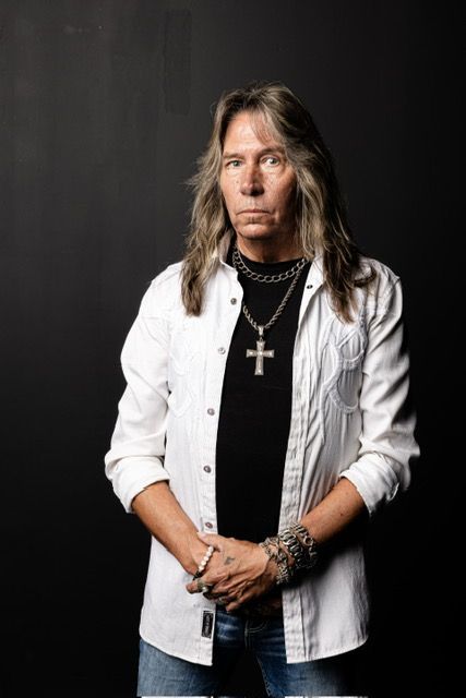 Man with long hair, wearing a white shirt over a black shirt, jeans, and a cross necklace.