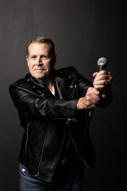 Man in a leather jacket holding a microphone, posing against a dark backdrop.