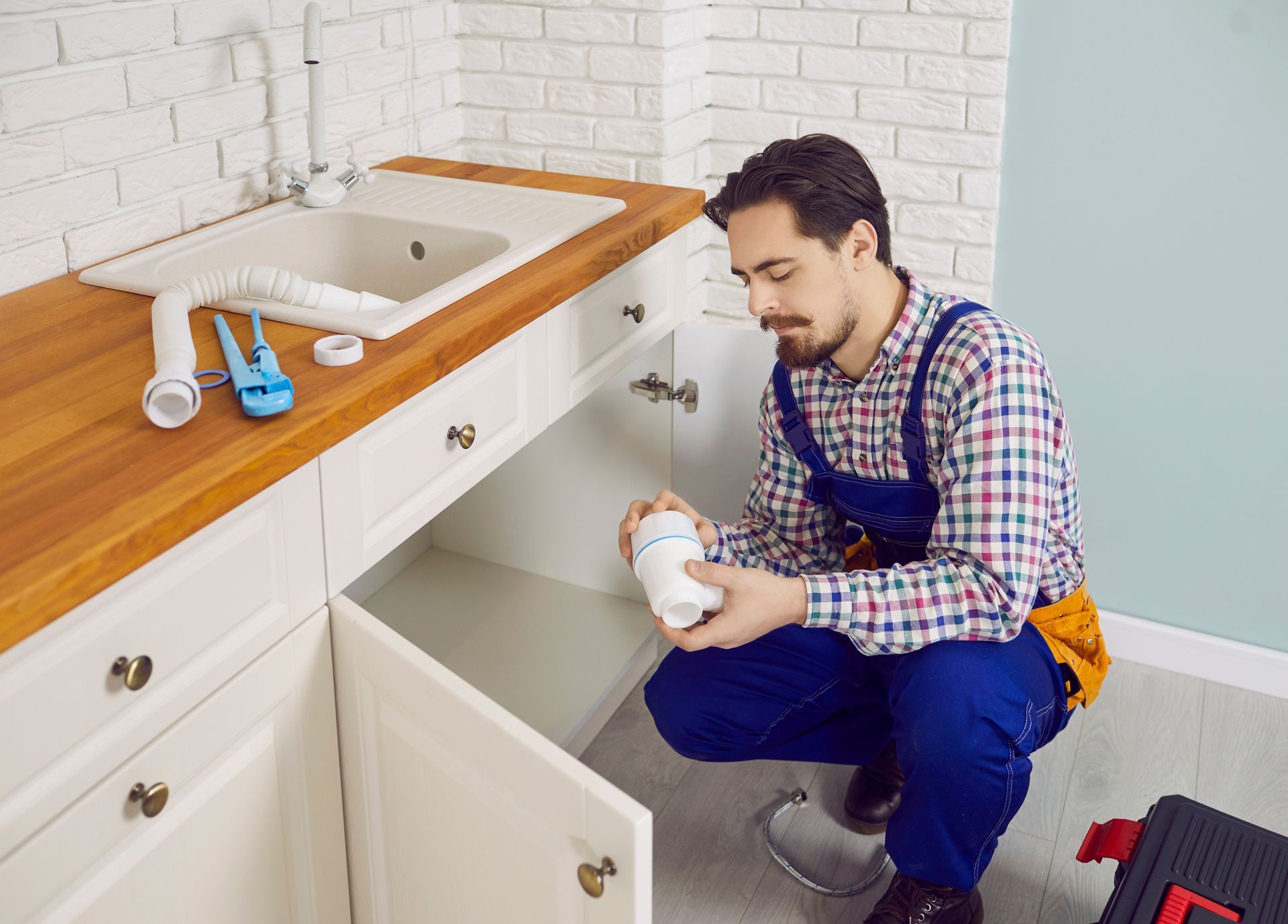 A plumbing company technician in blue overalls checks a drain trap by an open kitchen sink cabinet.