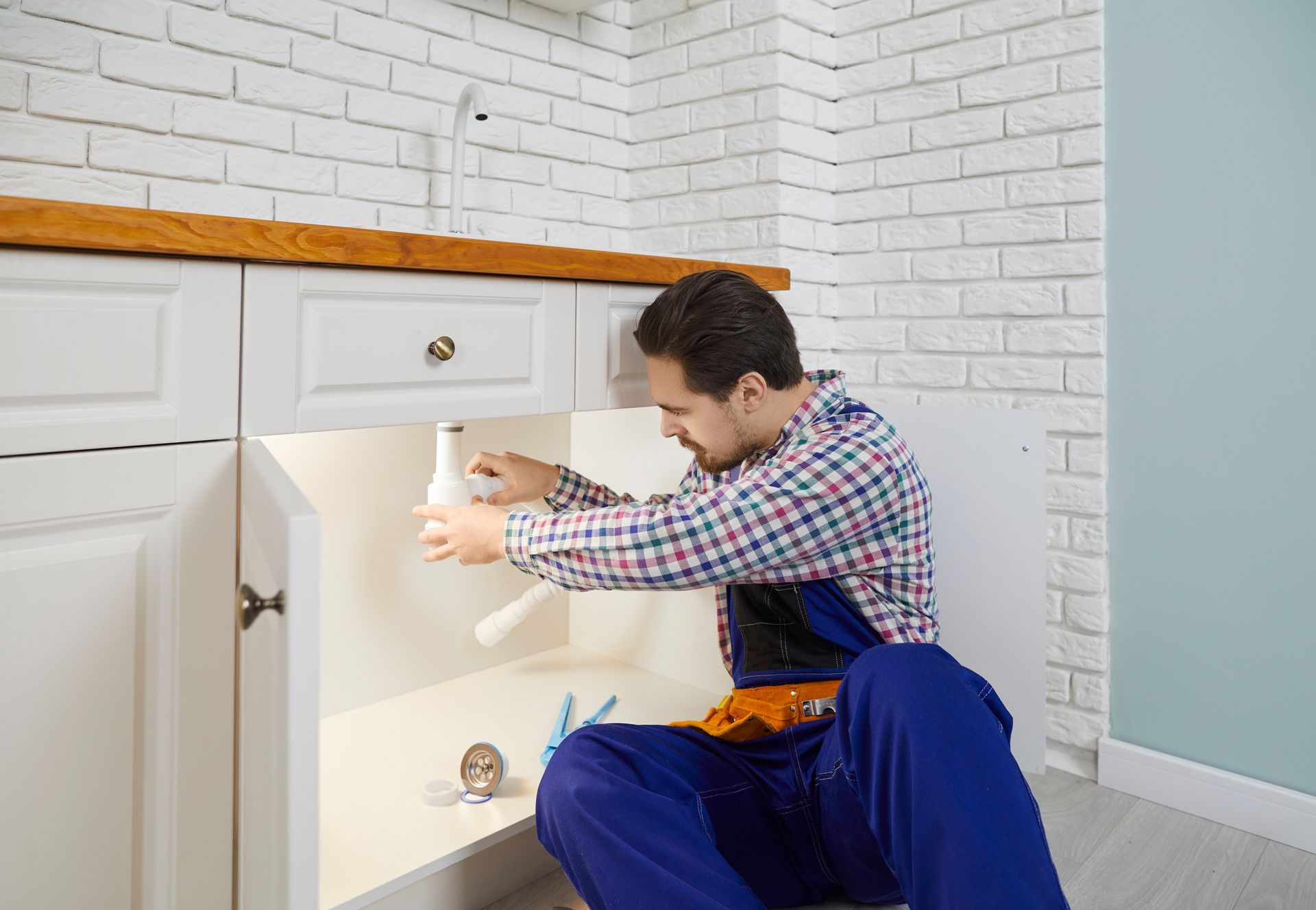 A plumbing company worker in blue overalls fixing a sink drain pipe under a white kitchen counter.
