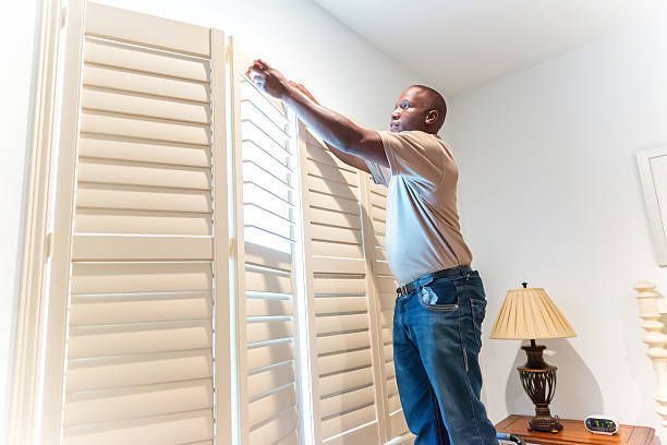 Man installing white plantation shutters on large window in bright living room.