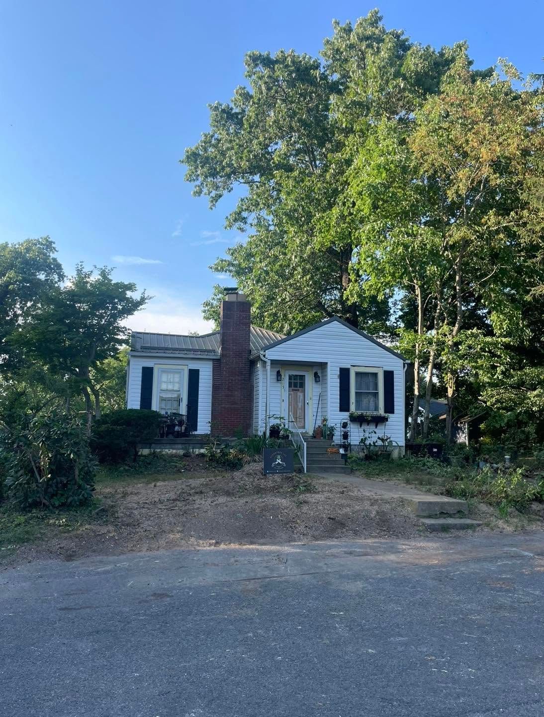 Small white house with dark shutters and red brick chimney, surrounded by trees along a roadside.