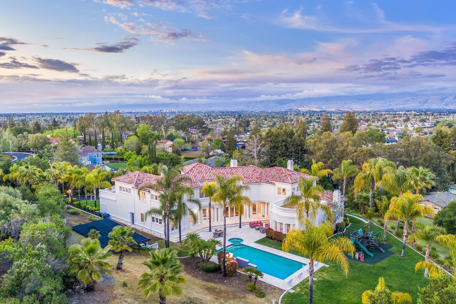 An aerial view of a large white house with a swimming pool surrounded by palm trees.