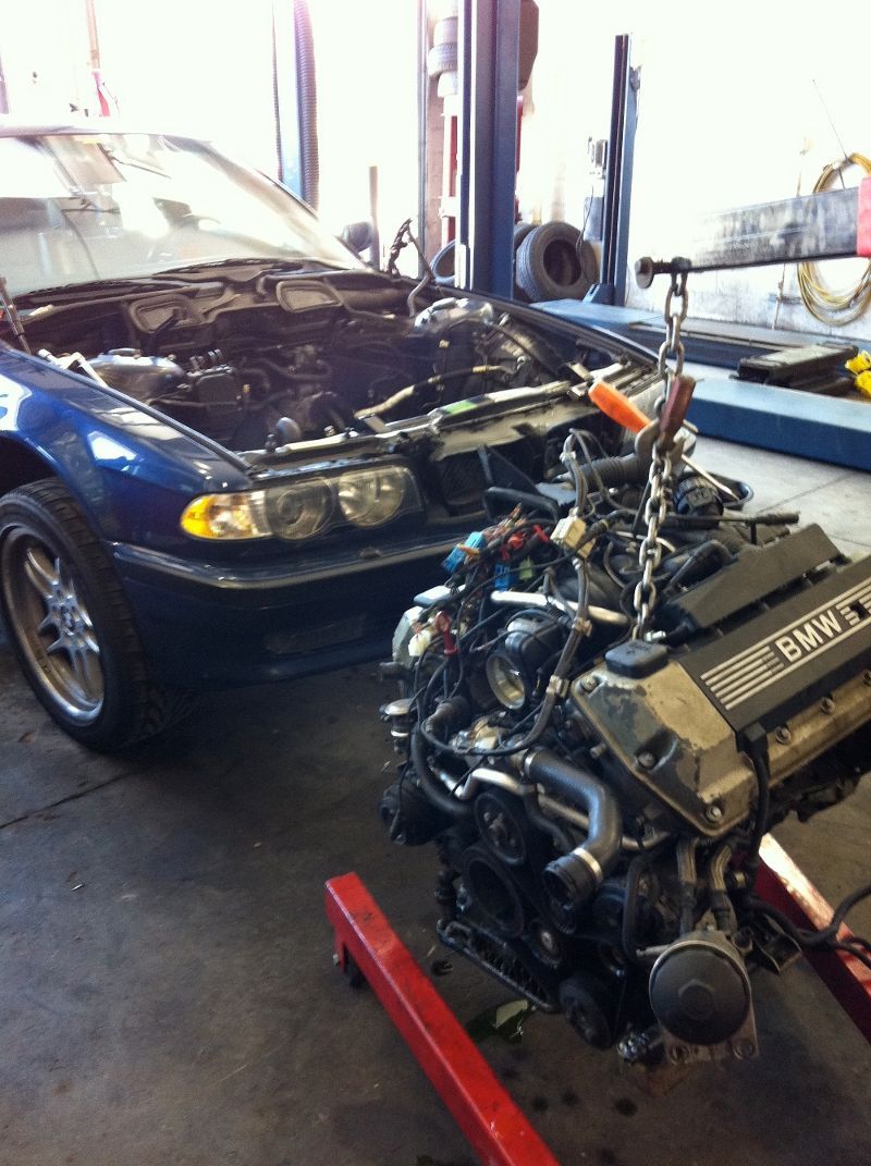 Blue sedan in a repair shop with its hood open, facing a partially assembled engine on a red stand.