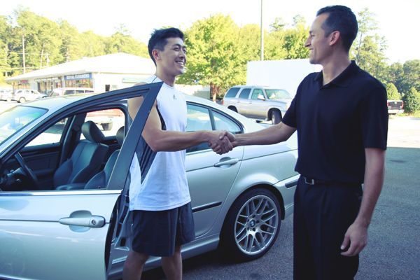 Two men shake hands beside a silver car in a parking lot.