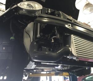 Black car on a lift with front bumper removed, exposing headlight and grille in a garage