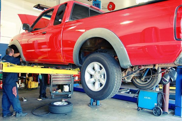 Red pickup truck raised on a lift in an auto repair shop, with a mechanic working underneath.