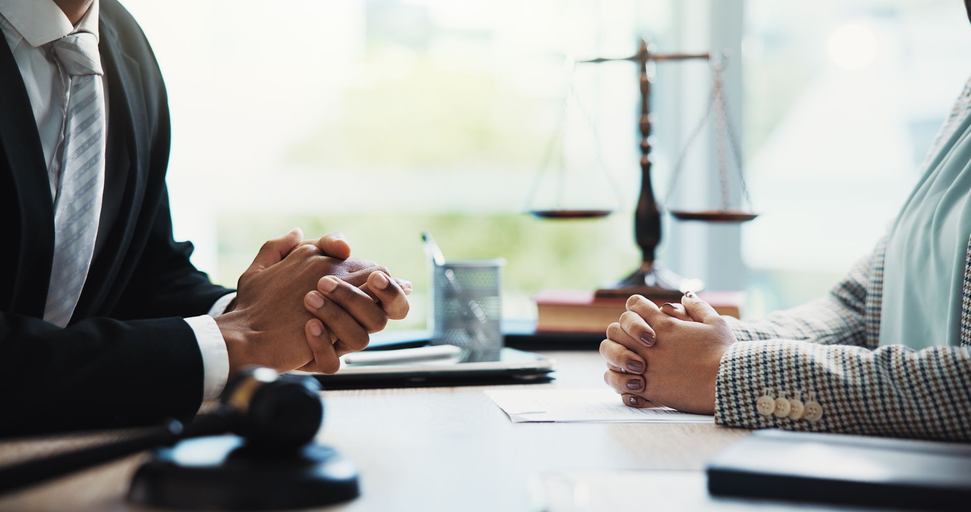 Two people at a desk, possibly lawyers, with clasped hands, scales of justice visible.