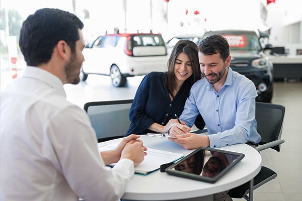 Car salesman discussing paperwork with a smiling couple at a desk in a bright showroom