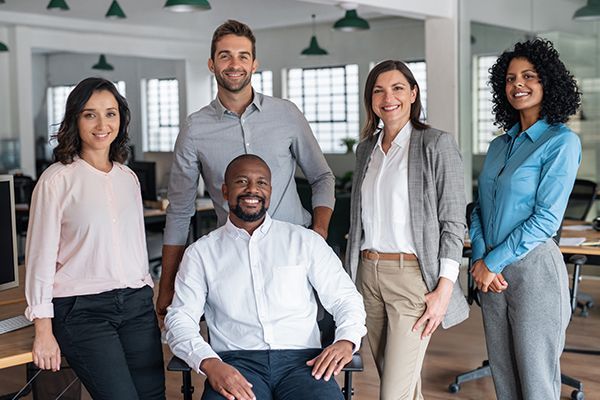 Five coworkers posing in a modern office, smiling at the camera with desks and hanging lights in the background