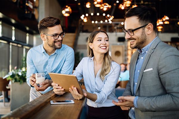 Three coworkers smiling and talking over coffee in a modern café while reviewing a tablet