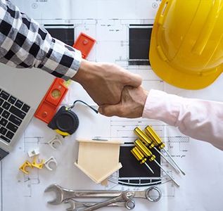 Two people shake hands over a desk with blueprints, tools, and a yellow hard hat, symbolizing a construction agreement