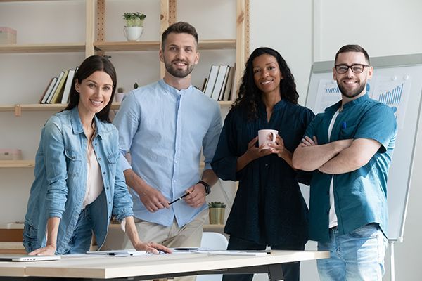 Four coworkers smiling in a bright office, standing together around a desk with laptops and papers.