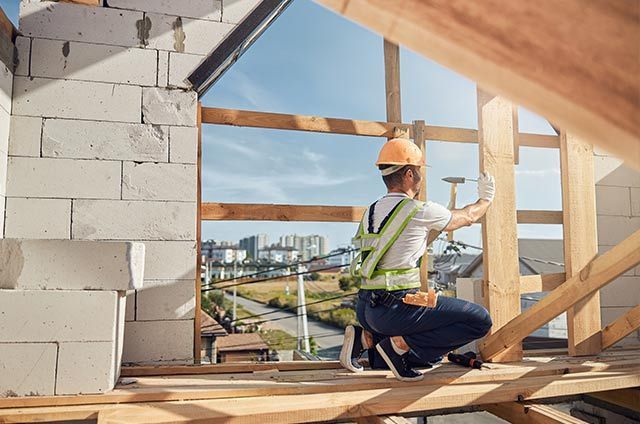Construction worker kneeling on a wooden frame, inspecting a house under construction with blue sky beyond.