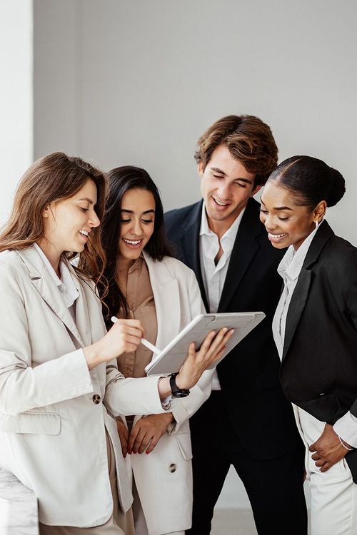 Four coworkers in light suits smiling while looking at a tablet in an office.