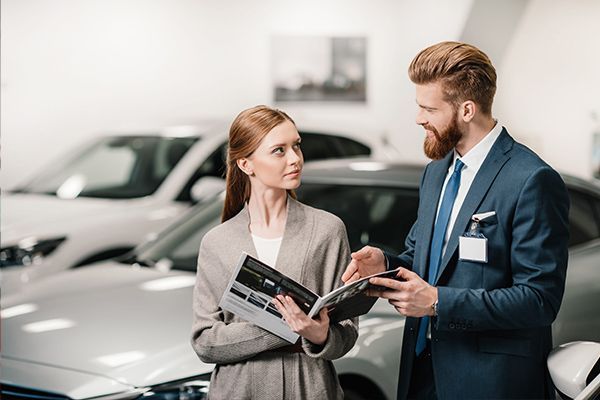 Two car salespeople reviewing brochures in a bright showroom with silver cars in the background