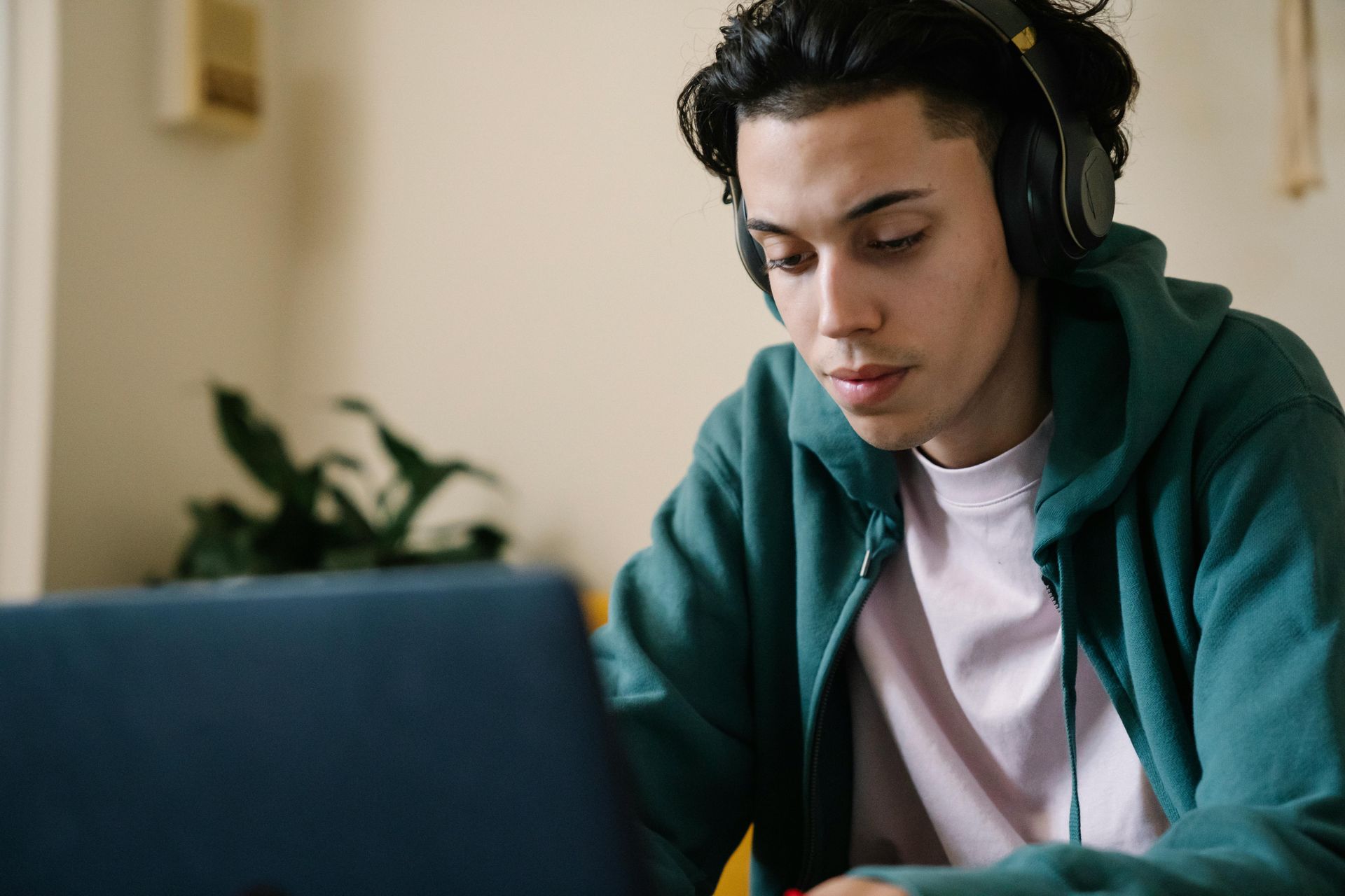 Student in a green hoodie wearing headphones, focused on a laptop in a bright room.
