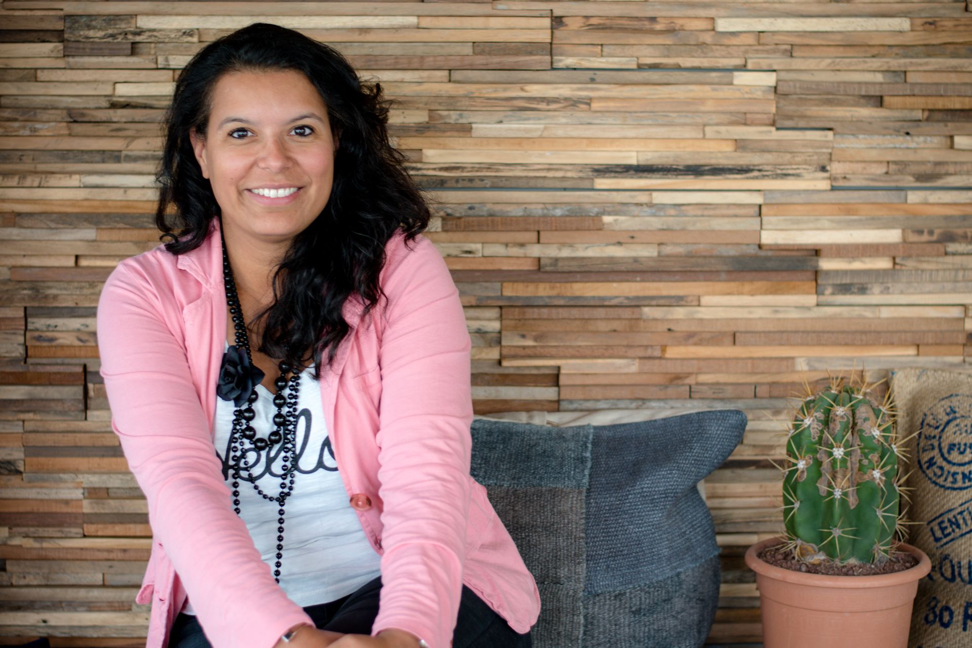 Woman in pink blazer smiles, seated by wooden wall, next to a cactus.