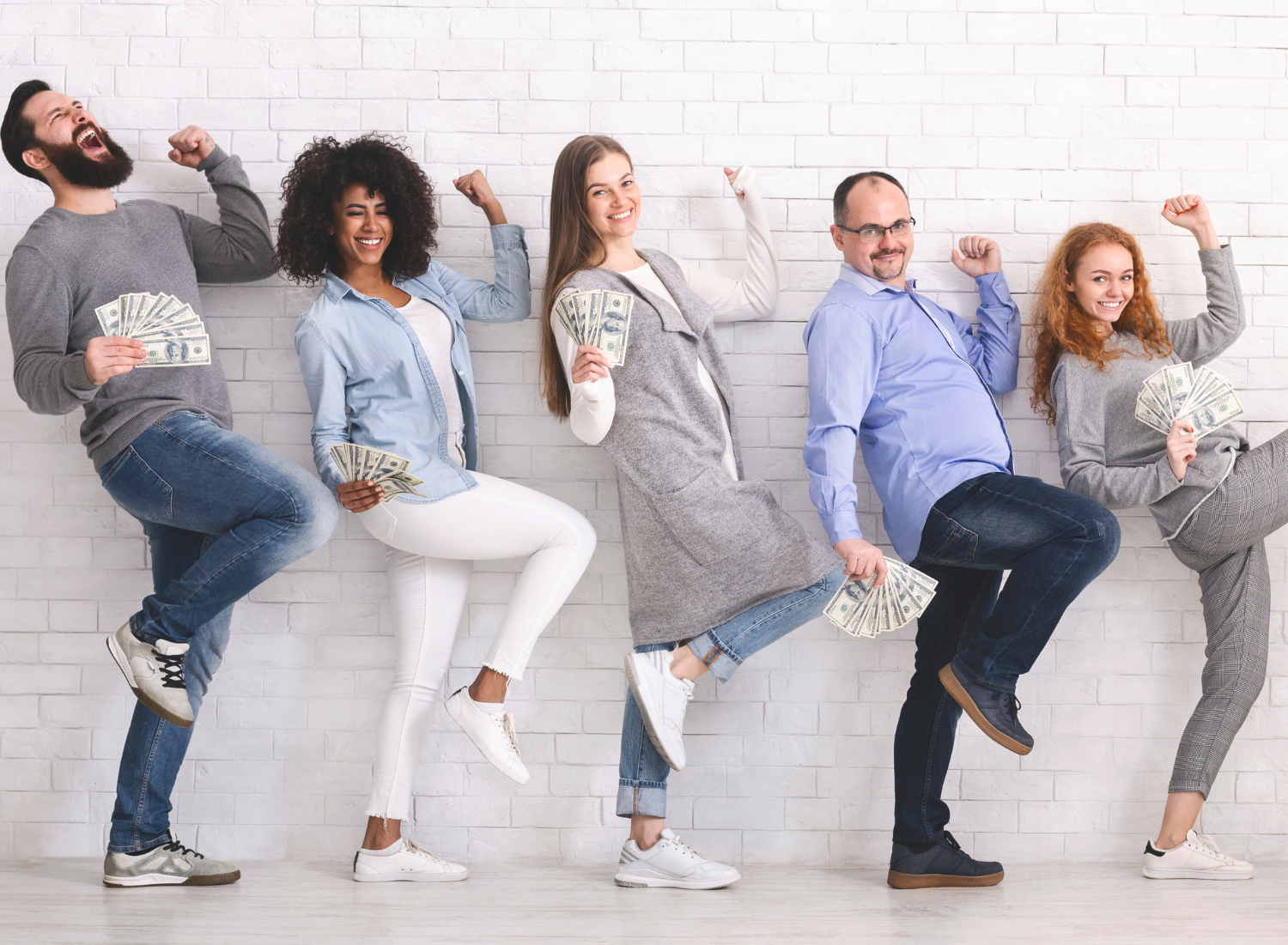 Five people celebrating with cash against a white brick wall. Smiling, happy expressions, arms raised.