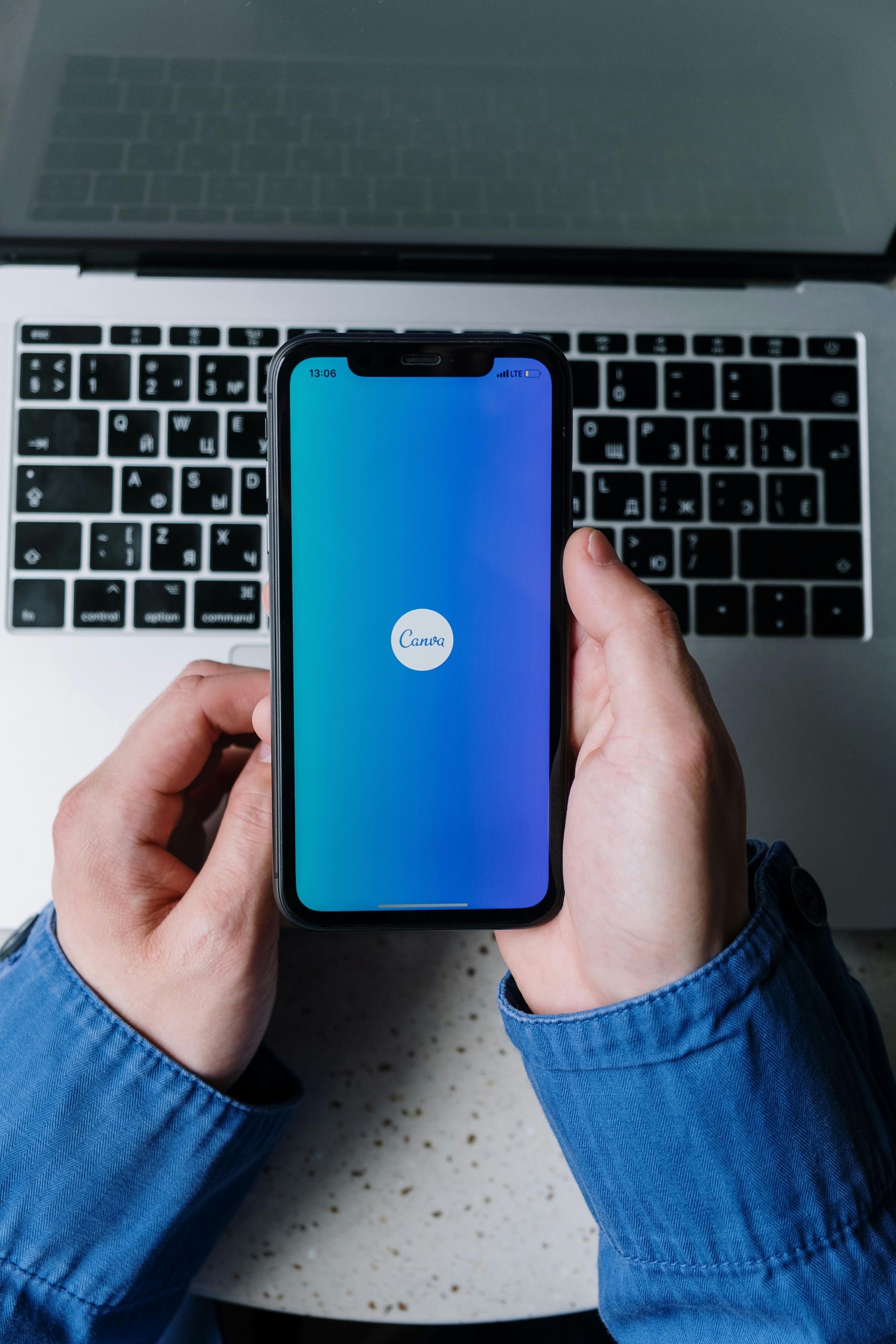 Hands holding phone displaying the Canva app on a blue-gradient background, with a laptop in the background.