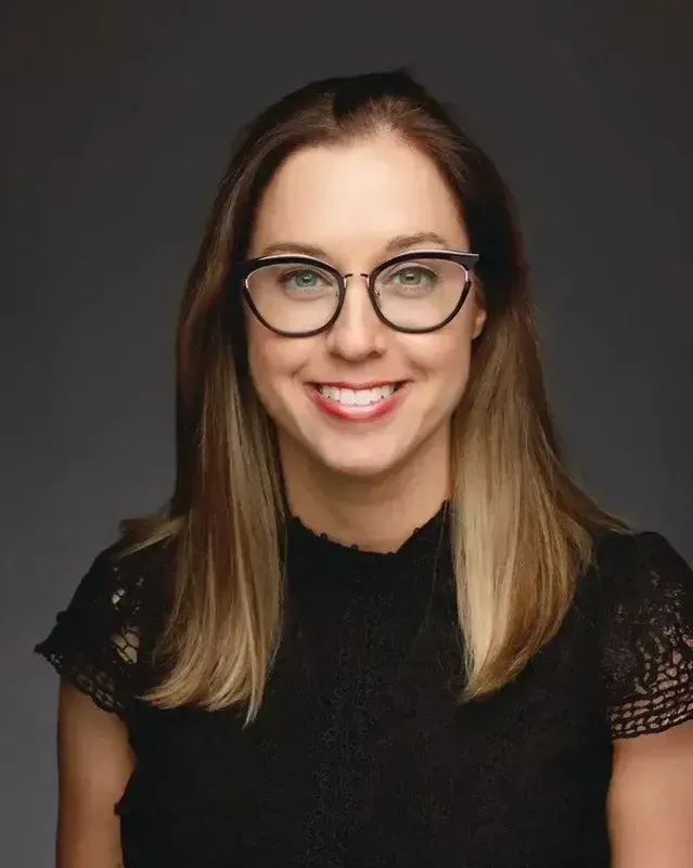 Studio headshot of a smiling person with glasses wearing a black top on a dark background