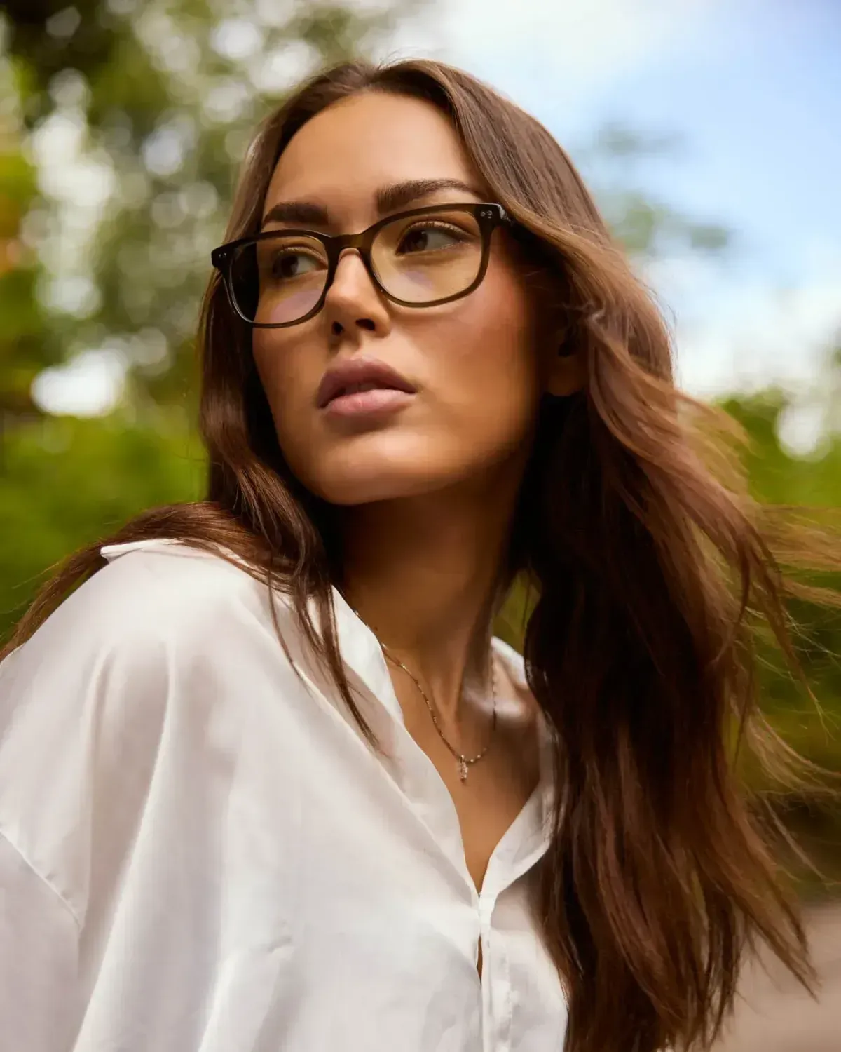Woman with glasses in a white shirt, looking off to the side outdoors with blurred greenery behind her