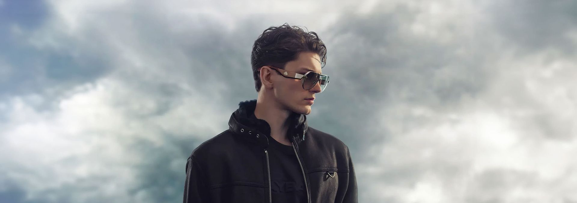 Person in sunglasses and black jacket standing against a cloudy sky.