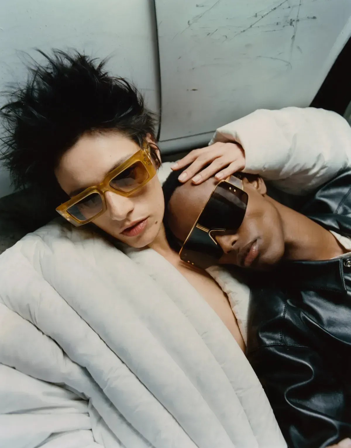 Two people reclining together in sunglasses and coats, one in a white puffer and one in black leather.