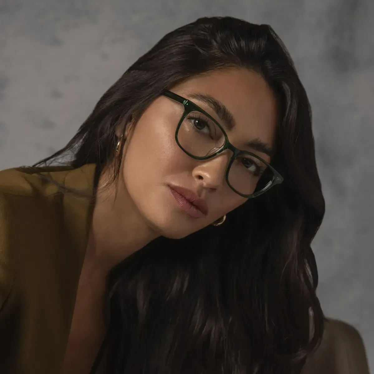 Woman with long dark hair and glasses posing against a gray studio backdrop.