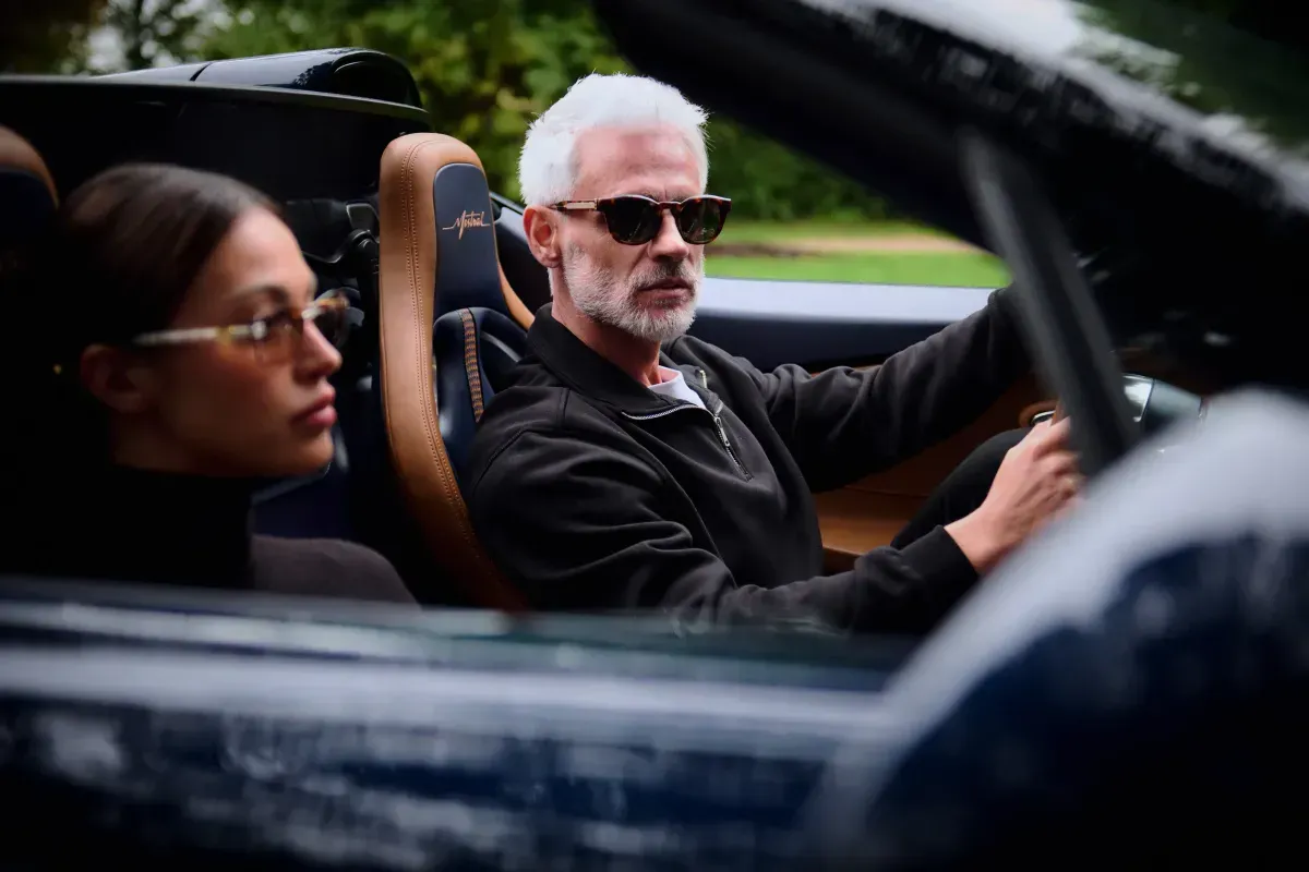 Two people riding in a convertible, one driving and one in profile wearing glasses.