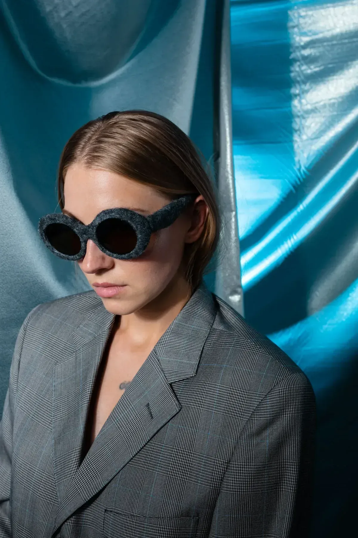 Person in round sunglasses and striped blazer against a shiny blue backdrop.