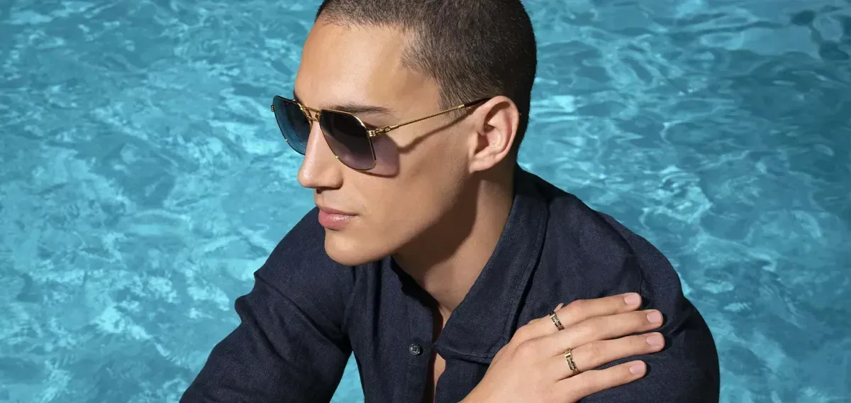 Person wearing sunglasses in a dark shirt, leaning by a turquoise pool with arms crossed.