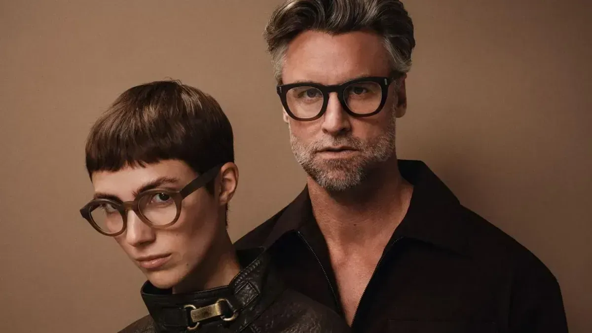 Two people wearing glasses against a brown background, posing closely together with neutral expressions.