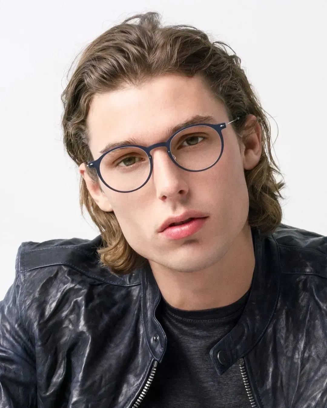 Young person with round glasses and wavy hair wearing a dark leather jacket against a light background
