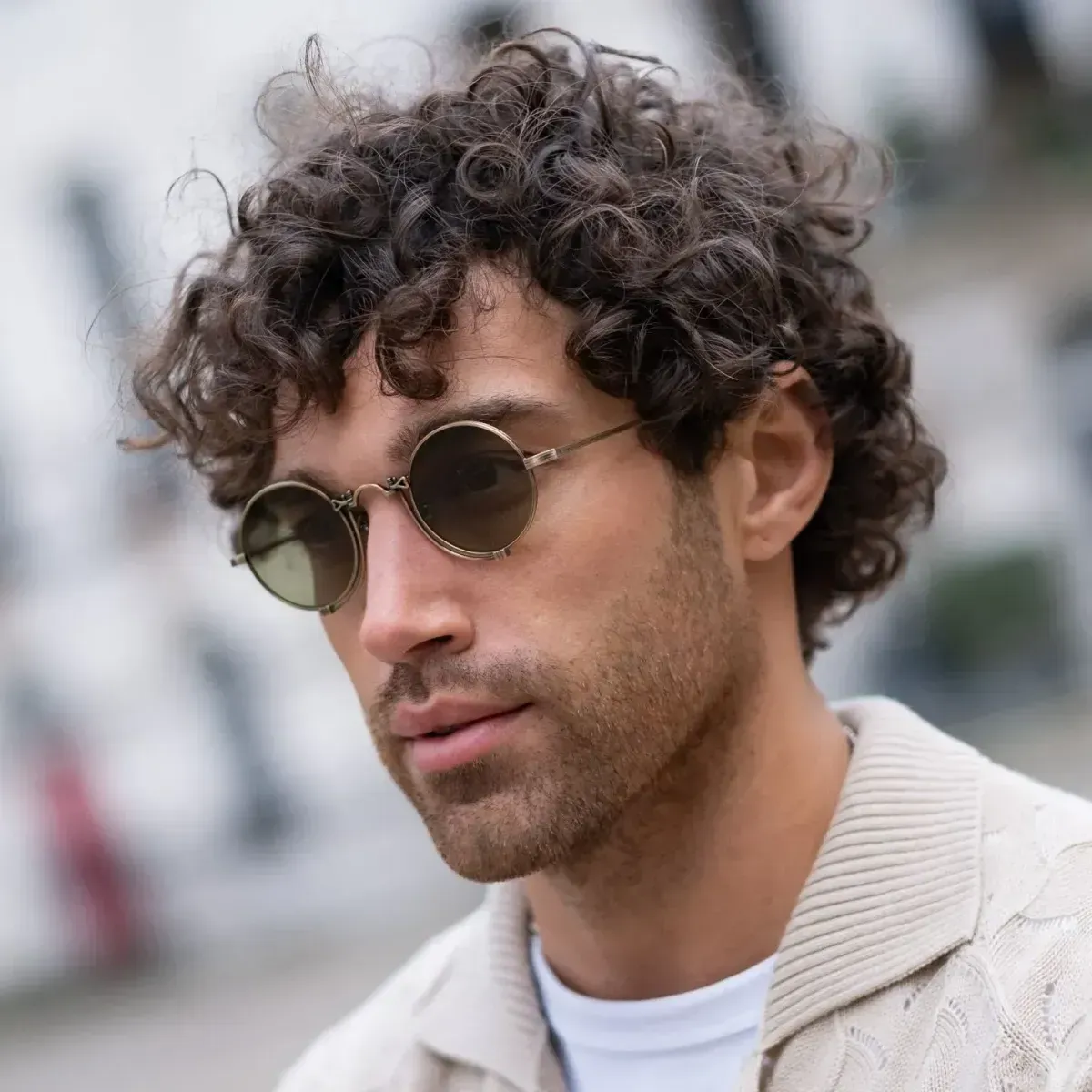 Close-up of a man with curly hair and round sunglasses, wearing a light sweater outdoors.